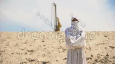 穿着白色衣服<strong>的</strong>阿拉伯人站在<strong>沙漠里</strong>，背景是石油井架，燃料生意